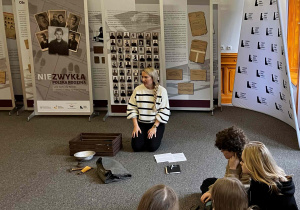 Na zdjęciu znajduje się prowadząca warsztaty Pani Patrycja Czyszpak-Pawlak, artefakty, uczniowie klasy 2c i w tle wystawa „Niezykła Polska Rodzina”.