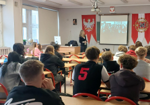 Szkolna klasa, uczniowie siedzą w ławkach, oglądają prezentację multimedialną